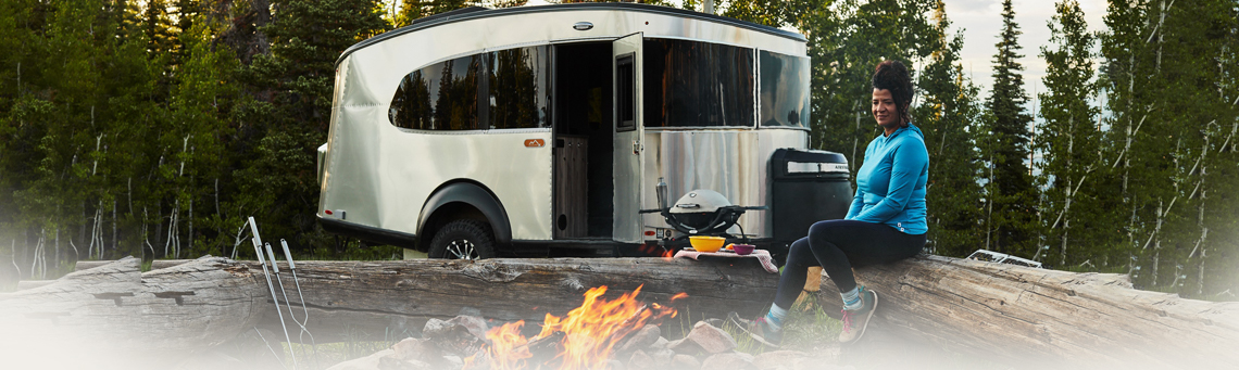 Woman sitting on a log near a campfire with an Airstream® RV parked in the background